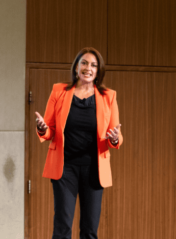 A woman wearing an orange blazer over black top and pants is standing on a stage speaking to an unseen audience. 